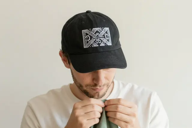 A Polish, White man wearing a black baseball cap with intricate white embroidery of a stylized, abstract pattern. The cap is positioned centrally in the frame, with a clean, neutral background that emphasizes the detailed stitching. The lighting highlights the texture and dimensionality of the embroidery.Art direction: Clean approachable lifestyle, dominant color palette: warm neutrals: off-white, cream, soft beige, sage, warm grey, with one gentle accent, accent color: green, light: soft natural daylight, airy and even, gentle shadows, low contrast, composition and framing: clean, balanced, human-centered, comfortable negative space, candid framing, finish/texture: soft, natural, organic textures, matte, no heavy filters, mood: welcoming,clear,genuine,approachable,calm. Style prompt: clean approachable lifestyle photography, soft natural daylight, warm neutral palette, balanced uncluttered composition, candid authentic moment, friendly and reassuring, shallow depth of field, sharp focus, 50mm, close-up on detailed embroidery, hands crafting textiles, personalized apparel, custom hats, corporate branding, unique gifts, high-quality stitching
