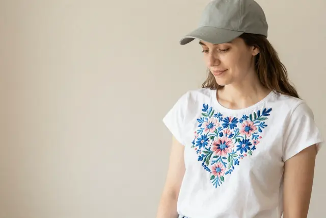 A Polish white woman in her late 20s proudly displays a custom-embroidered white t-shirt featuring a vibrant, intricate floral design in shades of blue and pink, a testament to personalized textile craftsmanship.Art direction: Clean approachable lifestyle, dominant color palette: warm neutrals: off-white, cream, soft beige, sage, warm grey, with one gentle accent, accent color: green, light: soft natural daylight, airy and even, gentle shadows, low contrast, composition and framing: clean, balanced, human-centered, comfortable negative space, candid framing, finish/texture: soft, natural, organic textures, matte, no heavy filters, mood: welcoming,clear,genuine,approachable,calm. Style prompt: clean approachable lifestyle photography, soft natural daylight, warm neutral palette, balanced uncluttered composition, candid authentic moment, friendly and reassuring, shallow depth of field, sharp focus, 50mm, close-up on detailed embroidery, hands crafting textiles, personalized apparel, custom hats, corporate branding, unique gifts, high-quality stitching