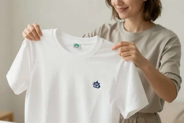 A Polish woman, white, with a friendly expression, is holding up a white t-shirt with a precisely embroidered navy blue logo on the chest. The t-shirt is neatly folded, showcasing the quality of the embroidery. The background is a clean, bright studio setting with soft lighting.Art direction: Clean approachable lifestyle, dominant color palette: warm neutrals: off-white, cream, soft beige, sage, warm grey, with one gentle accent, accent color: green, light: soft natural daylight, airy and even, gentle shadows, low contrast, composition and framing: clean, balanced, human-centered, comfortable negative space, candid framing, finish/texture: soft, natural, organic textures, matte, no heavy filters, mood: welcoming,clear,genuine,approachable,calm. Style prompt: clean approachable lifestyle photography, soft natural daylight, warm neutral palette, balanced uncluttered composition, candid authentic moment, friendly and reassuring, shallow depth of field, sharp focus, 50mm, close-up on detailed embroidery, hands crafting textiles, personalized apparel, custom hats, corporate branding, unique gifts, high-quality stitching